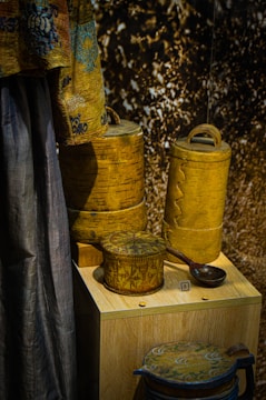 A display of intricately designed wooden containers and a fabric drape. The containers have detailed carvings and patterns, showcasing expert craftsmanship. The drape features a decorative design with blue and brown tones. The background has a textured appearance.
