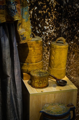A display of intricately designed wooden containers and a fabric drape. The containers have detailed carvings and patterns, showcasing expert craftsmanship. The drape features a decorative design with blue and brown tones. The background has a textured appearance.