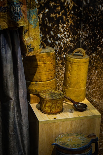 A display of intricately designed wooden containers and a fabric drape. The containers have detailed carvings and patterns, showcasing expert craftsmanship. The drape features a decorative design with blue and brown tones. The background has a textured appearance.