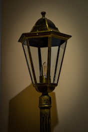 A vintage-style lamp post with a transparent glass enclosure housing an LED bulb, set against a plain wall. The lamp is designed with an ornate top and a weathered finish, casting a soft shadow.