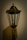 A vintage-style lamp post with a transparent glass enclosure housing an LED bulb, set against a plain wall. The lamp is designed with an ornate top and a weathered finish, casting a soft shadow.
