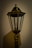 A vintage-style lamp post with a transparent glass enclosure housing an LED bulb, set against a plain wall. The lamp is designed with an ornate top and a weathered finish, casting a soft shadow.
