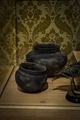 Two ancient-looking black pottery vessels are displayed on a wooden surface, adjacent to a decorative bird sculpture. The background features an intricately patterned wallpaper in muted tones.