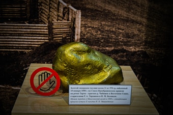 A large, shiny golden nugget is displayed on a wooden plinth, accompanied by a sign in Russian and a red circle symbol with a hand, indicating 'do not touch'. The background features a dark, textured surface that contrasts with the gleaming nugget.