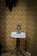 A small, round table covered with a delicately crocheted lace doily holds an antique oil lamp. The table stands against a wall adorned with vintage, ornate floral wallpaper in gold and beige tones.