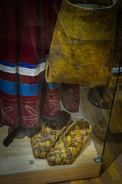 A pair of woven shoes and a birch bark container are displayed on a wooden surface inside a glass case. The shoes have a detailed basket-like pattern, and the container shows natural textures. The background features red fabric adorned with intricate white embroidery and blue accents.