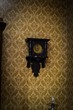 An ornate antique wall clock with a dark wooden frame and a yellow clock face. The background features a richly patterned mustard-colored wallpaper with floral designs. Below the clock, there is a glass oil lamp with a tall, narrow chimney.