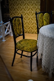 A vintage-style chair with a dark wooden frame and green patterned upholstery is positioned next to a round table draped with a lace tablecloth. The chair features intricate floral designs in the fabric. The room has a wooden floor and an ornate wallpaper with a similar floral pattern in shades of brown and gold, adding to the antique ambiance.