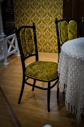 A vintage-style chair with a dark wooden frame and green patterned upholstery is positioned next to a round table draped with a lace tablecloth. The chair features intricate floral designs in the fabric. The room has a wooden floor and an ornate wallpaper with a similar floral pattern in shades of brown and gold, adding to the antique ambiance.