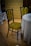 A vintage-style chair with a dark wooden frame and green patterned upholstery is positioned next to a round table draped with a lace tablecloth. The chair features intricate floral designs in the fabric. The room has a wooden floor and an ornate wallpaper with a similar floral pattern in shades of brown and gold, adding to the antique ambiance.