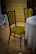 A vintage-style chair with a dark wooden frame and green patterned upholstery is positioned next to a round table draped with a lace tablecloth. The chair features intricate floral designs in the fabric. The room has a wooden floor and an ornate wallpaper with a similar floral pattern in shades of brown and gold, adding to the antique ambiance.