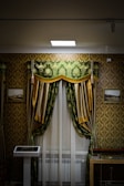 An ornately decorated room features a window with luxurious green and gold curtains, white sheer drapes, and a decorative pelmet. The walls are covered in a gold and brown damask wallpaper. There are framed pictures on either side of the window, and a display case is partially visible in the lower right corner. A modern touch screen display stands to the left of the window.