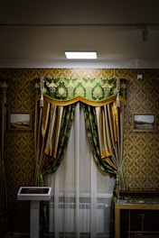 An ornately decorated room features a window with luxurious green and gold curtains, white sheer drapes, and a decorative pelmet. The walls are covered in a gold and brown damask wallpaper. There are framed pictures on either side of the window, and a display case is partially visible in the lower right corner. A modern touch screen display stands to the left of the window.