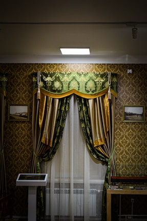 An ornately decorated room features a window with luxurious green and gold curtains, white sheer drapes, and a decorative pelmet. The walls are covered in a gold and brown damask wallpaper. There are framed pictures on either side of the window, and a display case is partially visible in the lower right corner. A modern touch screen display stands to the left of the window.