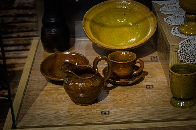 A collection of brown ceramic pottery displayed in a glass case, including cups, bowls, and jugs, arranged neatly on wooden platforms with numbered labels. White decorative doilies are partially visible in the background.