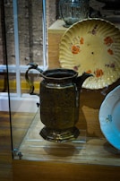 A vintage display featuring a brass pitcher with intricate carvings, a decorative floral plate in yellowish tones, and a glass container in the background. These items are showcased in a glass case on a wooden base.