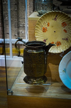 A vintage display featuring a brass pitcher with intricate carvings, a decorative floral plate in yellowish tones, and a glass container in the background. These items are showcased in a glass case on a wooden base.