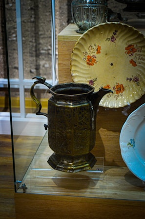 A vintage display featuring a brass pitcher with intricate carvings, a decorative floral plate in yellowish tones, and a glass container in the background. These items are showcased in a glass case on a wooden base.