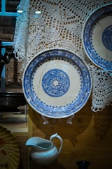 A display featuring intricately patterned blue and white ceramic plates, one prominently showcased on a stand. The background includes a delicate white lace fabric. Below the plates, a white ceramic pitcher with a blue rim is visible. The setting appears to be a curated collection, possibly in a museum or antique shop.