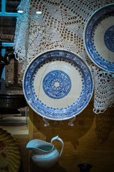 A display featuring intricately patterned blue and white ceramic plates, one prominently showcased on a stand. The background includes a delicate white lace fabric. Below the plates, a white ceramic pitcher with a blue rim is visible. The setting appears to be a curated collection, possibly in a museum or antique shop.