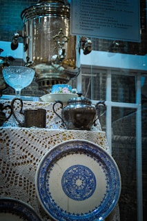 A collection of vintage kitchenware items displayed together. A large brass samovar takes a prominent position atop a delicately woven lace tablecloth. Surrounding it are an assortment of objects including an ornate crystal bowl, a silver teapot, and a blue-patterned ceramic plate. Some small metal cups and a floral-patterned cup and saucer add to the eclectic assortment. A sign with text in Russian is visible at the top.