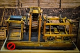 A wooden mechanical model featuring gears, pulleys, and rollers is displayed against a sepia-toned background that resembles industrial or construction settings. The structure includes a conveyor system and a rotating drum with holes. The wood is stained in different shades, giving it a vintage appearance.