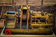 A wooden mechanical model featuring gears, pulleys, and rollers is displayed against a sepia-toned background that resembles industrial or construction settings. The structure includes a conveyor system and a rotating drum with holes. The wood is stained in different shades, giving it a vintage appearance.