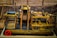 A wooden mechanical model featuring gears, pulleys, and rollers is displayed against a sepia-toned background that resembles industrial or construction settings. The structure includes a conveyor system and a rotating drum with holes. The wood is stained in different shades, giving it a vintage appearance.