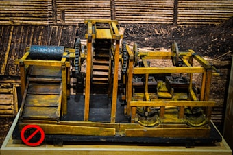 A wooden mechanical model featuring gears, pulleys, and rollers is displayed against a sepia-toned background that resembles industrial or construction settings. The structure includes a conveyor system and a rotating drum with holes. The wood is stained in different shades, giving it a vintage appearance.
