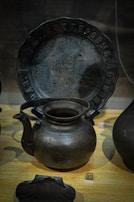 A collection of dark metal artifacts is displayed on a wooden surface. A notable piece is a large, round metal plate with inscriptions along the rim. In front of the plate, a rustic metal kettle with a curved spout and large handle is prominently featured. To the side, there is a number plaque labeled 17. The artifacts exhibit an ancient or vintage appearance.