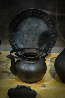 A collection of dark metal artifacts is displayed on a wooden surface. A notable piece is a large, round metal plate with inscriptions along the rim. In front of the plate, a rustic metal kettle with a curved spout and large handle is prominently featured. To the side, there is a number plaque labeled 17. The artifacts exhibit an ancient or vintage appearance.