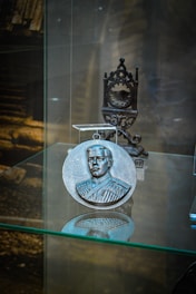A round metallic medallion with an embossed portrait of a man in a formal uniform is displayed on a glass shelf. Behind the medallion, an intricately designed metal stand is visible. The background is blurred, with hints of a wooden structure.