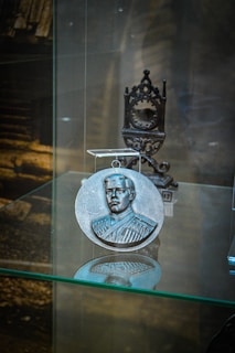 A round metallic medallion with an embossed portrait of a man in a formal uniform is displayed on a glass shelf. Behind the medallion, an intricately designed metal stand is visible. The background is blurred, with hints of a wooden structure.