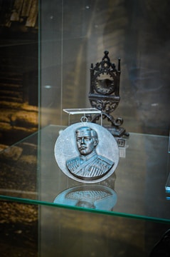 A round metallic medallion with an embossed portrait of a man in a formal uniform is displayed on a glass shelf. Behind the medallion, an intricately designed metal stand is visible. The background is blurred, with hints of a wooden structure.