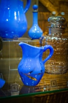 A collection of glassware is displayed on a shelf. The foreground features a blue ceramic pitcher with a delicate handle and a gold leaf design. There are also other blue and clear glass containers, such as decanters and bottles, in the background. Each item on the shelf is accompanied by a numbered label.