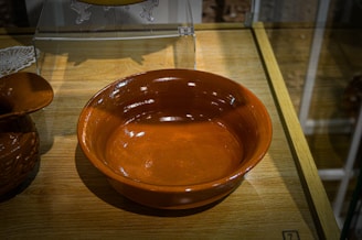 A glazed ceramic bowl with a rich terracotta color is displayed on a wooden surface. The bowl's reflective surface captures light, highlighting its smooth texture. There is an intricate ceramic item partially visible to the left, suggesting a similar craftsmanship.