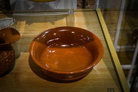 A glazed ceramic bowl with a rich terracotta color is displayed on a wooden surface. The bowl's reflective surface captures light, highlighting its smooth texture. There is an intricate ceramic item partially visible to the left, suggesting a similar craftsmanship.