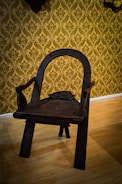 An intricately carved wooden chair with a dark finish, featuring a curved backrest and sturdy legs. The chair is positioned on a light-colored wooden floor with a background of ornate, gold damask wallpaper.