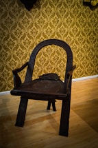 An intricately carved wooden chair with a dark finish, featuring a curved backrest and sturdy legs. The chair is positioned on a light-colored wooden floor with a background of ornate, gold damask wallpaper.