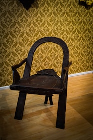 An intricately carved wooden chair with a dark finish, featuring a curved backrest and sturdy legs. The chair is positioned on a light-colored wooden floor with a background of ornate, gold damask wallpaper.