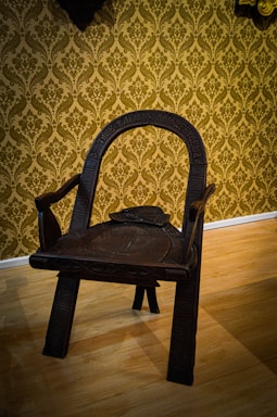 An intricately carved wooden chair with a dark finish, featuring a curved backrest and sturdy legs. The chair is positioned on a light-colored wooden floor with a background of ornate, gold damask wallpaper.