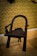 An intricately carved wooden chair with a dark finish, featuring a curved backrest and sturdy legs. The chair is positioned on a light-colored wooden floor with a background of ornate, gold damask wallpaper.