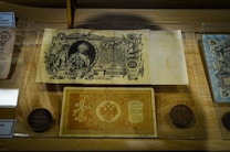 A display case featuring vintage banknotes and coins. The central note has detailed engravings and portraits, surrounded by ornate patterns. Coins with visible inscriptions are arranged nearby, all set on a wooden background.