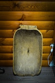 A large, aged wooden basin is positioned against a textured wooden wall with a warm brown finish. An electrical socket and a white cable with a switch are visible on the right side. The basin has visible wear and cracks, giving it a rustic and vintage appearance.