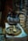 An old, blackened metal pot sits on a white cloth-covered surface alongside a large, rustic wooden dish holding two wooden utensils. Above the pot is a hook, and in the background, various objects and textures are visible, suggesting an antique or historical setting.