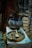An old, blackened metal pot sits on a white cloth-covered surface alongside a large, rustic wooden dish holding two wooden utensils. Above the pot is a hook, and in the background, various objects and textures are visible, suggesting an antique or historical setting.