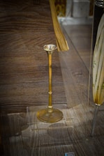 An antique brass object with a long stem and a flared top on display in a transparent case, placed on a wooden surface. There are additional objects partially visible around the brass piece, but the main focus is on the brass item.