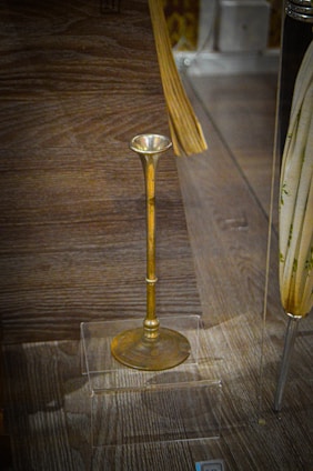 An antique brass object with a long stem and a flared top on display in a transparent case, placed on a wooden surface. There are additional objects partially visible around the brass piece, but the main focus is on the brass item.