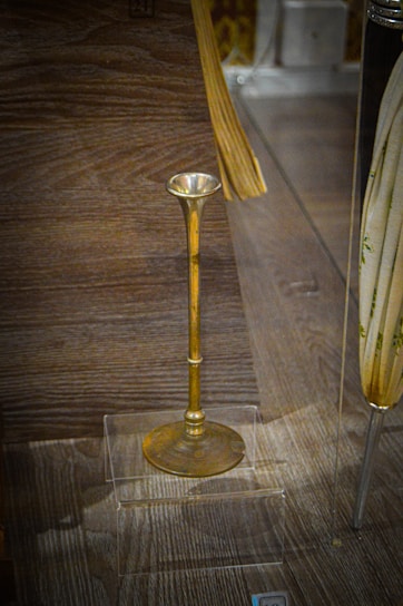 An antique brass object with a long stem and a flared top on display in a transparent case, placed on a wooden surface. There are additional objects partially visible around the brass piece, but the main focus is on the brass item.
