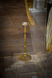 An antique brass object with a long stem and a flared top on display in a transparent case, placed on a wooden surface. There are additional objects partially visible around the brass piece, but the main focus is on the brass item.
